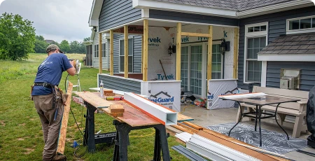 Carpenter renovating a house porch with boards on sawhorses and exposed framing.